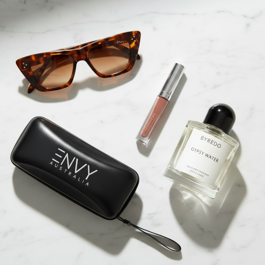 A styled flat-lay on a white marble surface featuring a black Envy Australia sunglasses case, tortoiseshell sunglasses with brown lenses, a tube of lipstick, and a bottle of Byredo Gypsy Water perfume.