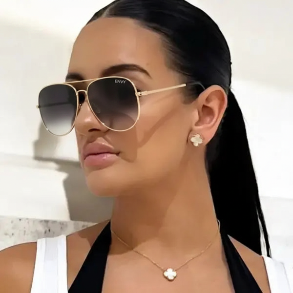 Woman wearing gold sunglasses with 'ENVY' branding, black hair tied back, and white top.