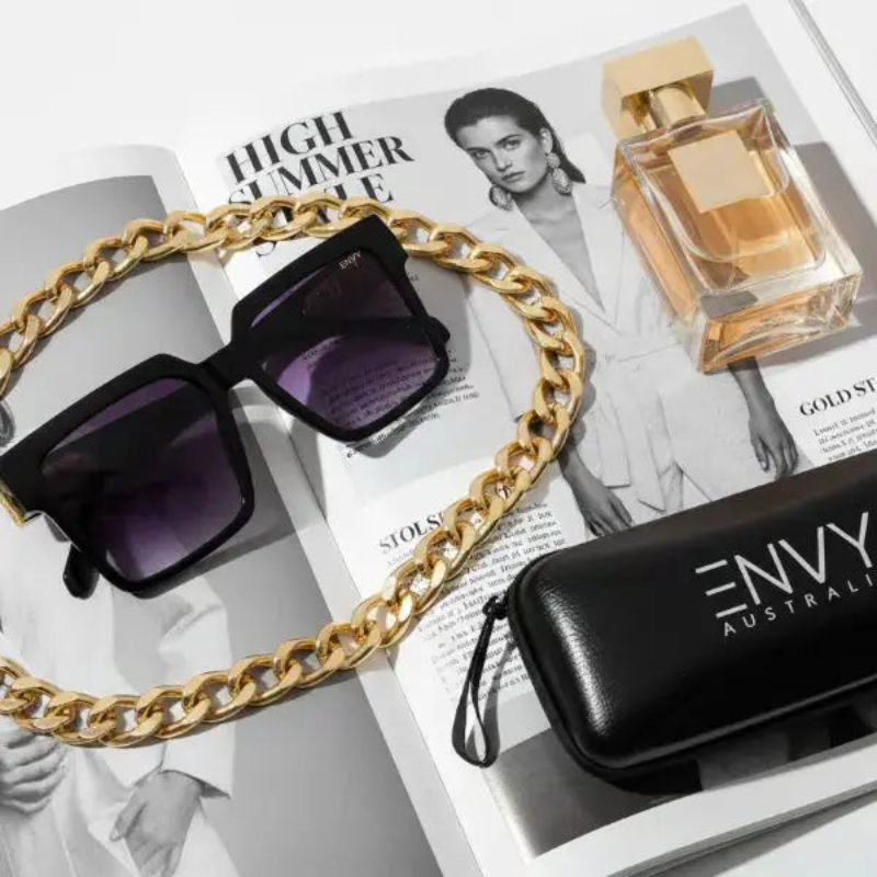 Sunglasses with gold chain, black case, and perfume bottle on a magazine