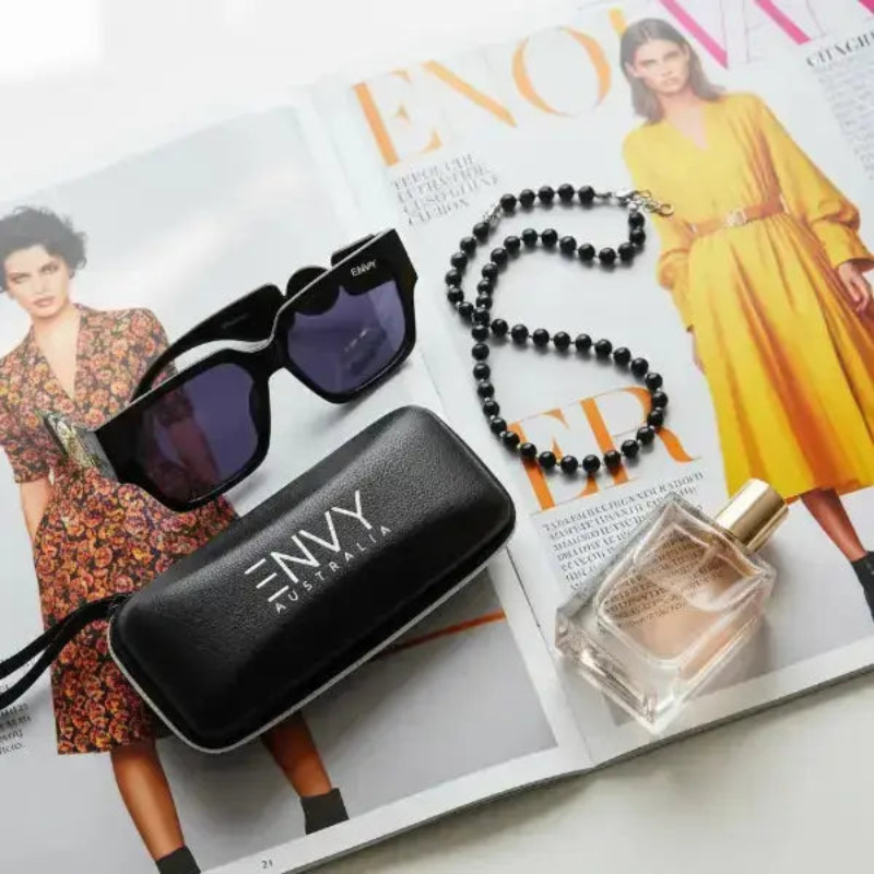 Black Milan sunglasses with case, black beaded necklace, and perfume bottle on an open magazine.