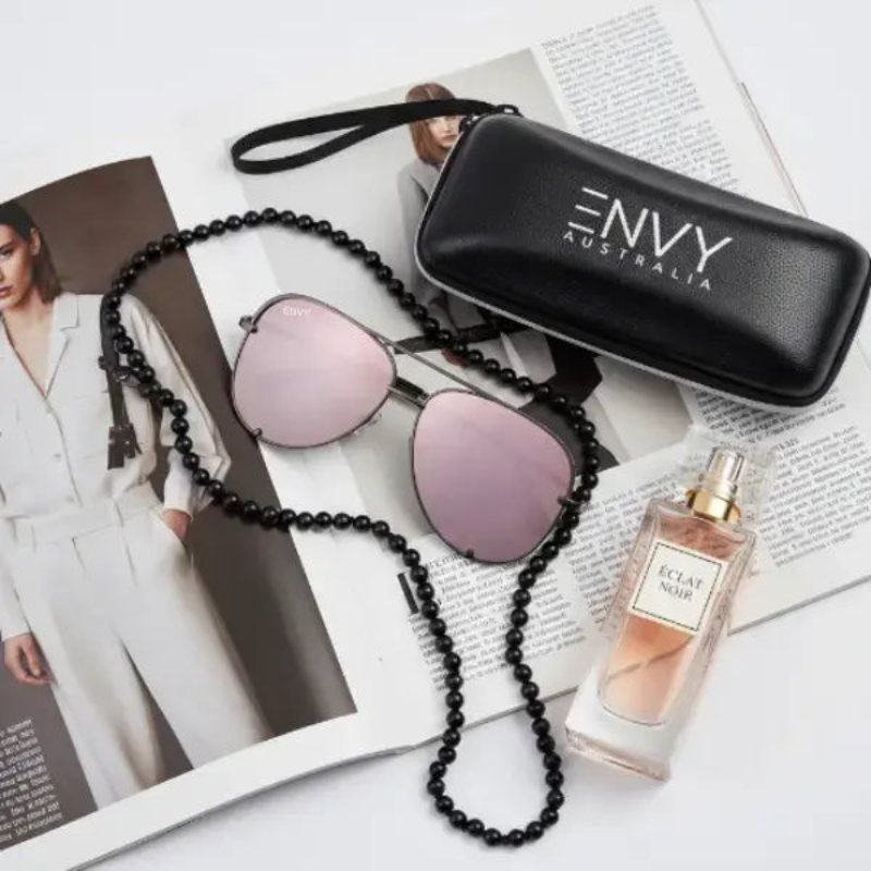 Mirror Shine Pink sunglasses with a beaded chain, Envy Australia case, and a perfume bottle on a magazine.