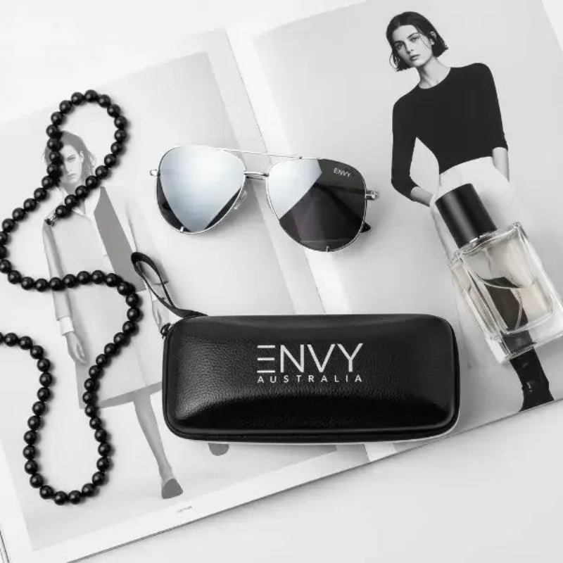 Black 'ENVY Australia' sunglasses case with Mirror Shine Silver sunglasses, necklace, and perfume bottle on a magazine