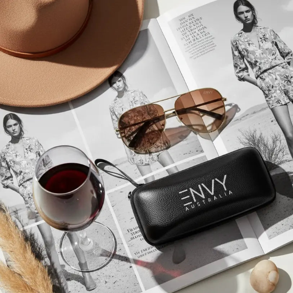 Sunglasses with 'ENVY Australia' case on a magazine with a glass of red wine and a hat.
