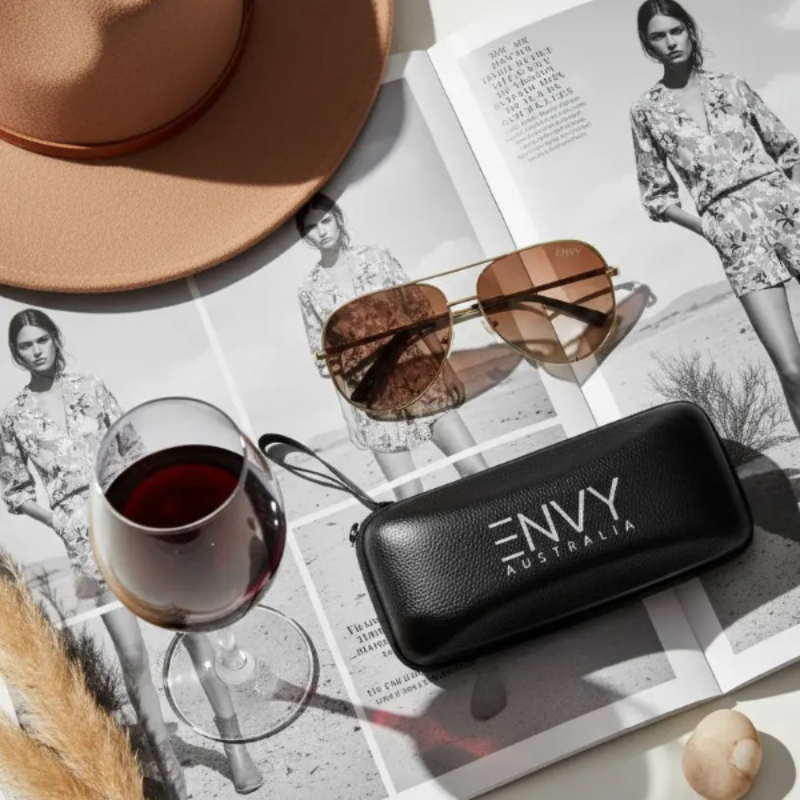 Ombre Pilot Tan aviator sunglasses with 'ENVY Australia' case on a magazine with a glass of red wine and a hat.