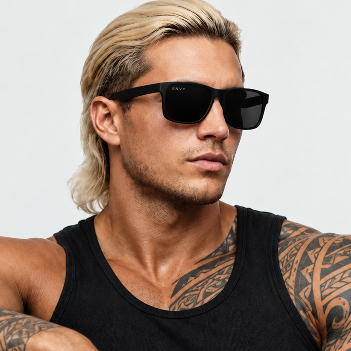 A man with blonde hair and tattoos wearing Onyx Matte black sunglasses and a black tank top, photographed against a light gray studio background.