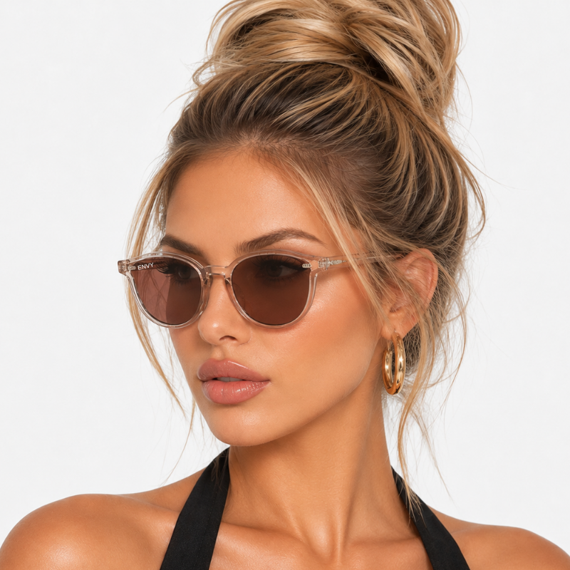 A close-up of a woman with blonde hair in a high bun wearing cat-eye sunglasses with a clear acetate frame and brown lenses, set against a plain white background.