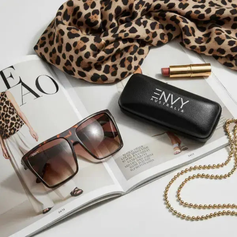 Sachi Sunglasses, leopard print scarf, and lip balm on an open magazine with 'ENVY Australia' case.