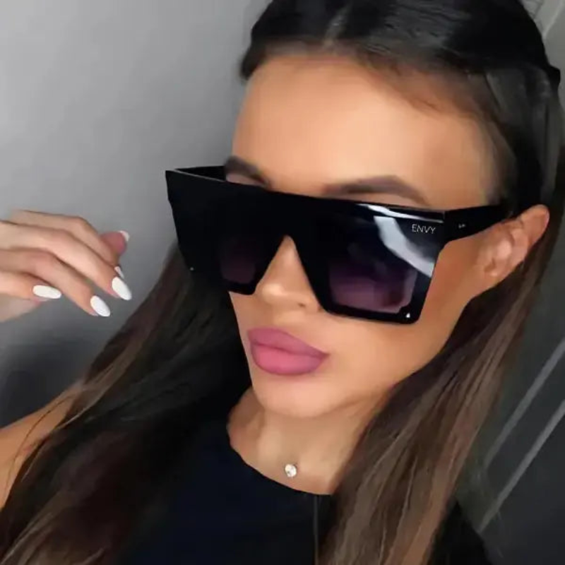 Woman wearing Selena Deluxe black sunglasses with 'ENVY' branding, indoors.