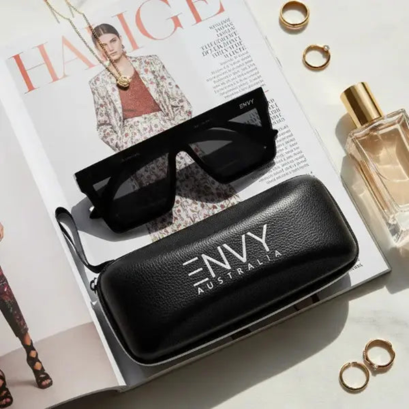 Selena Deluxe Black sunglasses with case and perfume bottle on a magazine