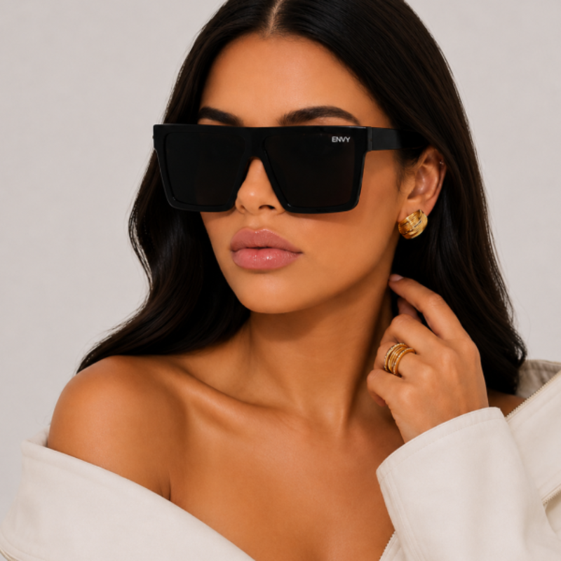 Woman wearing Selena Deluxe black sunglasses with 'ENVY' logo, gold earrings, and rings on a neutral background
