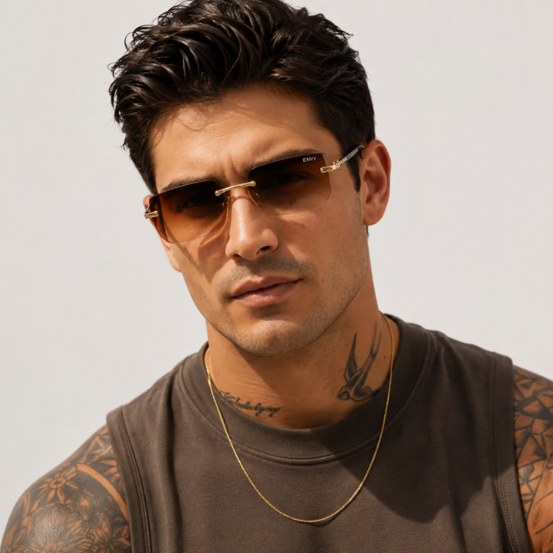 A close-up of a man wearing slim aviator-style sunglasses with gold metal frames and brown tinted lenses, paired with a brown sleeveless shirt and a gold chain necklace against a light gray background.