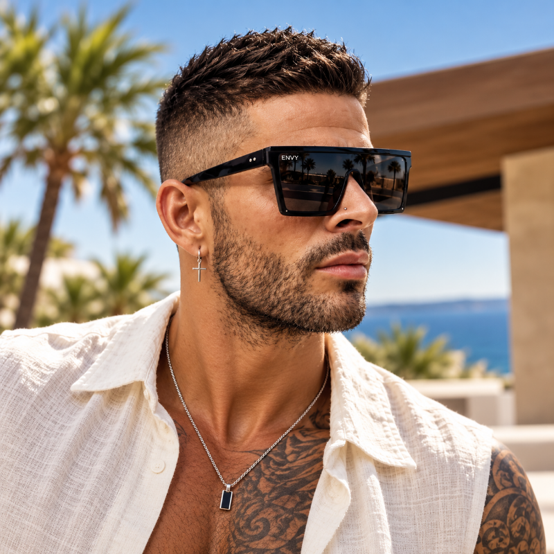 A man wearing Blade Black sunglasses with a small silver-toned logo on the frame, styled outdoors with palm trees and a blue sea in the background.