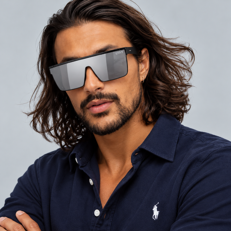 A man with long wavy brown hair and a beard wearing oversized shield-style sunglasses with a black frame and silver mirrored lenses, paired with a navy Polo Ralph Lauren button-up shirt against a light gray background.