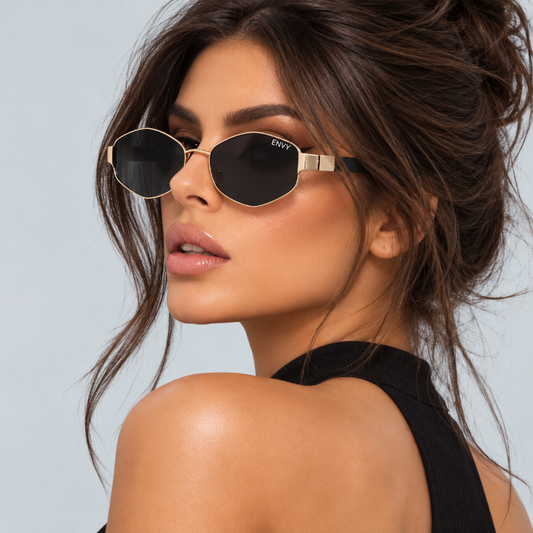A close-up of a woman wearing angular gold-framed sunglasses with dark lenses. The brand name 'ENVI' is visible on the temple arm.