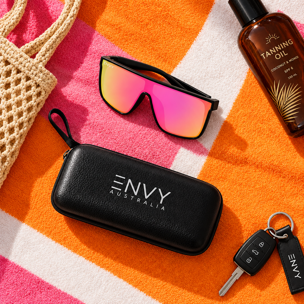 A black textured leather sunglasses case with 'ENVY AUSTRALIA' printed in white, styled in a flat lay on a pink, white, and orange striped towel alongside pink mirrored sunglasses, a bottle of tanning oil, a woven tote bag, and keys.