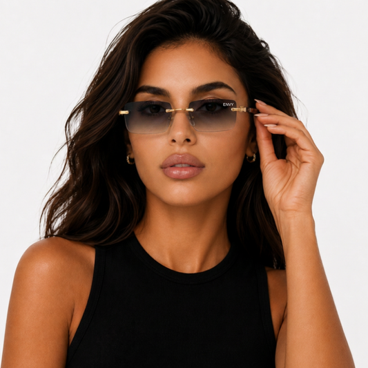A woman with dark hair wearing Envy Sunglasses rimless dark-tinted sunglasses with gold metal frames and temples, holding one lens with her hand against a plain white background.
