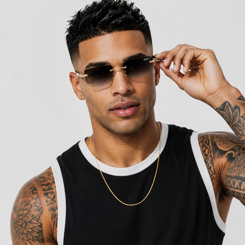 A man with tattoos wearing gold-framed sunglasses with dark lenses, a gold chain necklace, and a black sleeveless tank top with white trim, set against a plain light gray background.