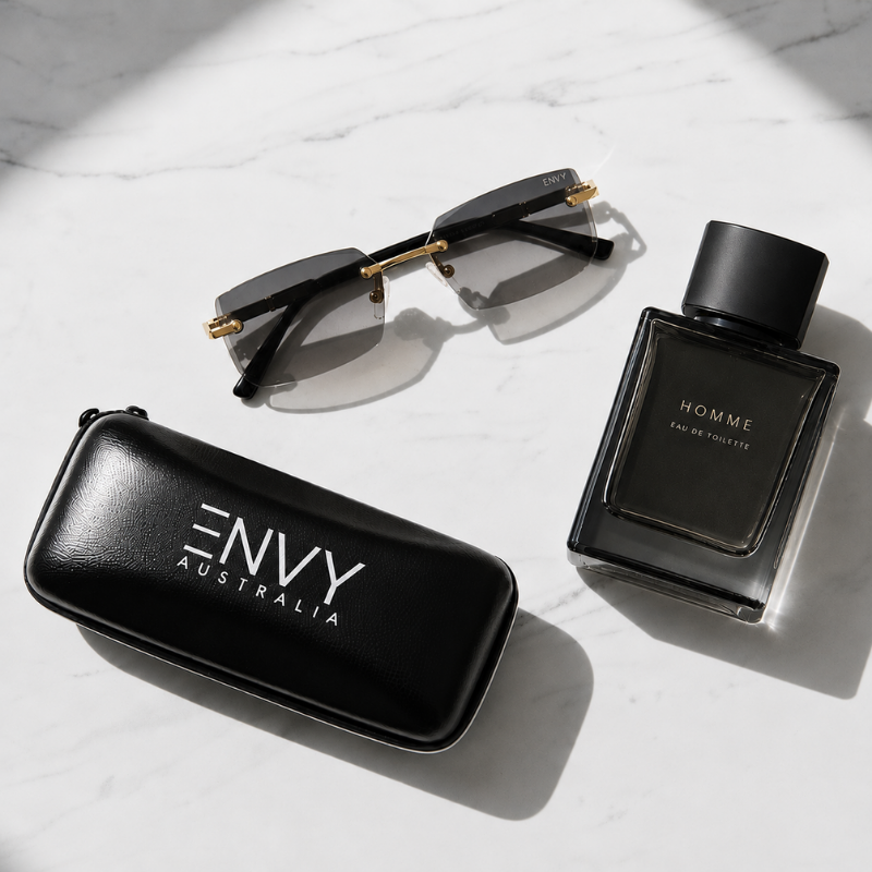 A luxury eyewear and fragrance set on a white marble background. The set includes a pair of black rimless sunglasses with gold metal temples, a black glass perfume bottle labeled 'HOMME', and a black leather case with 'ENVY AUSTRALIA' printed in white.