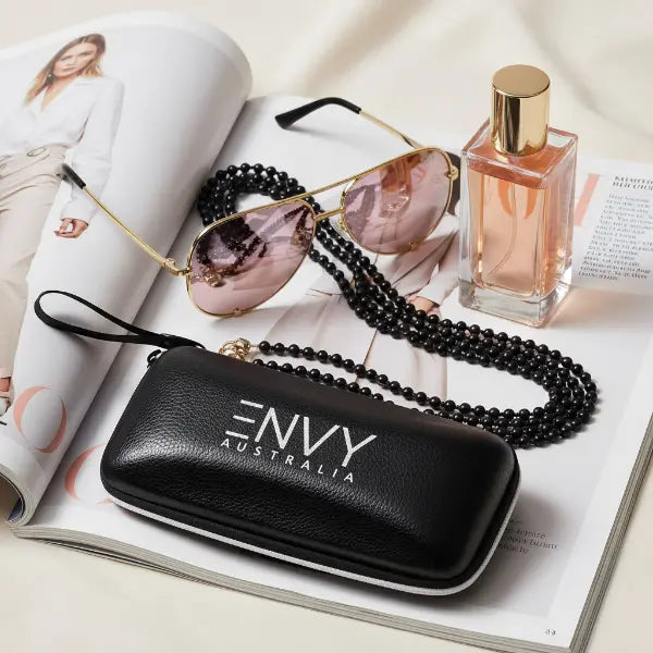 Black 'ENVY Australia' sunglasses case with sunglasses and a perfume bottle on an open magazine.
