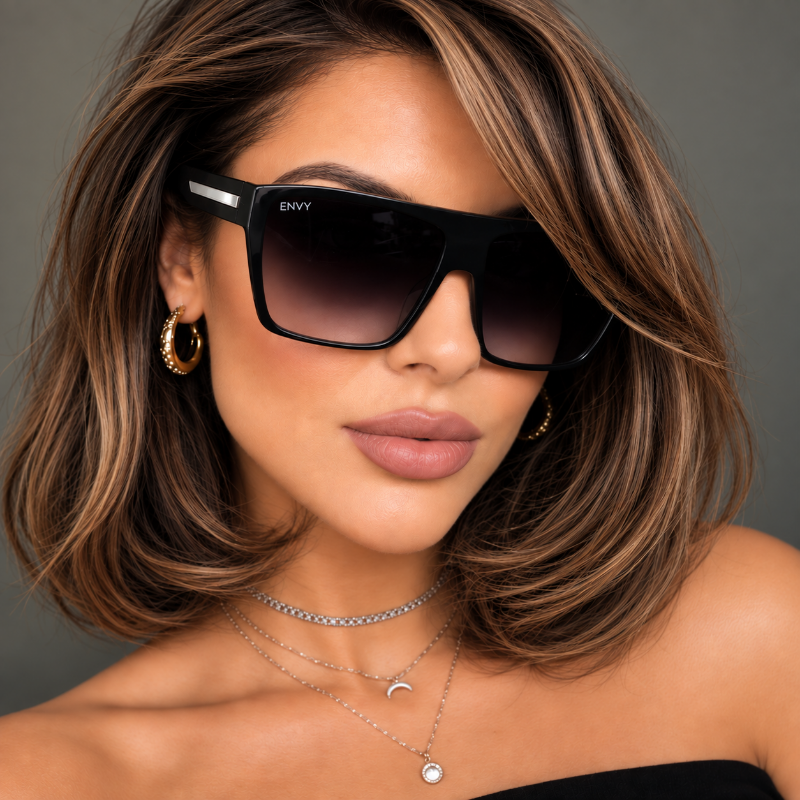 A close-up lifestyle shot of a woman with long brown hair wearing Sachi Oversized black sunglasses with a white accent stripe on the temple. The brand name 'ENVY' is visible on the upper corner of the lens.