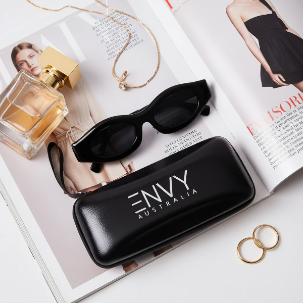 Black sunglasses with 'ENVY Australia' case on open magazine with perfume and jewelry.