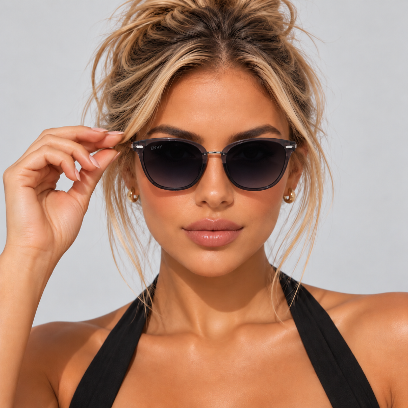 A close-up of a woman wearing black cat-eye sunglasses with dark lenses and thin metal frames, adjusting the side temple with her hand. She is wearing a black halter top against a light gray studio background.