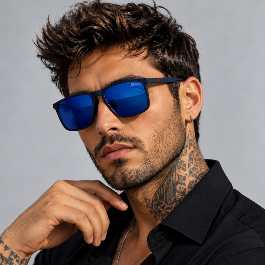 A close-up of a man wearing dark-framed sunglasses called Trader blue with vibrant blue mirrored lenses and a black shirt, set against a solid gray studio background.