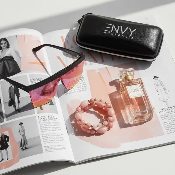 Black sunglasses with 'ENVY Australia' case on an open magazine with a bracelet and perfume bottle.
